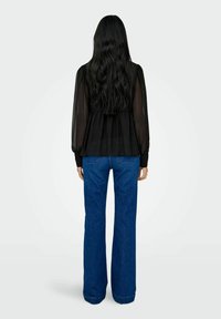 Femme aux longs cheveux noirs portant un chemisier noir transparent et un jean bleu taille haute, debout dos tourné vers l'avant.