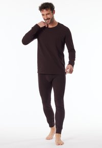 Kahvin pitkähihainen toppi ja kiinteät housut, jotka on valmistettu pehmeästä kankaasta. Topissa on pyöreä kaula-aukko ja saumaton muotoilu; malli seisoo paljain jaloin.