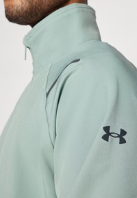 Svetle zelená sportovní bunda s vysokým límcem. Disponuje texturovanou tkaninou, černými akcenty a malým logem na rukávu.