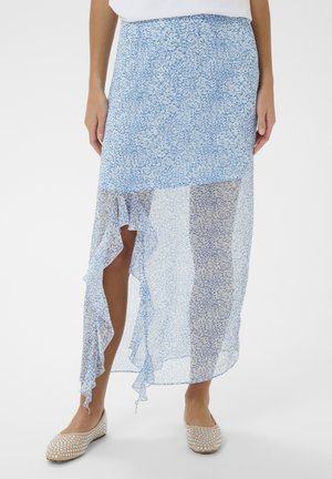 Žena v svetlomodrej midi sukni s kvetinovým vzorom, asymetrickým volánovým lemom a béžovými balerínkami so cvočkami.