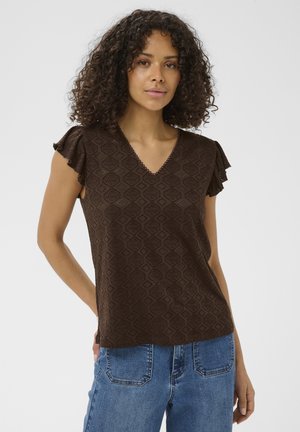 Femme aux cheveux bouclés portant un chemisier marron à motifs à manches courtes avec des volants aux manches et un jean en denim bleu, debout devant un fond uni.