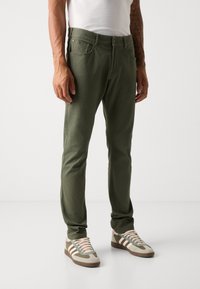 Pantalons en coton vert olive à coupe près du corps, avec poches avant et coutures discrètes. Associés à des baskets beiges et vert olive.