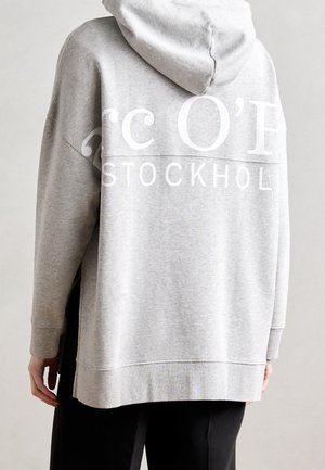 Person trägt einen hellgrauen, übergroßen Hoodie mit dem Schriftzug "STOCKHOLM" und teilweise großen weißen Buchstaben auf der Rückseite, kombiniert mit schwarzen Hosen.