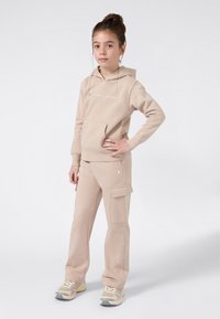 Jong meisje staat met haar hand op de heup, gekleed in een beige Champion hoodie, bijpassende cargo trainingsbroek en beige sneakers, tegen een effen witte achtergrond.