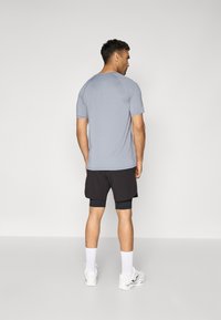 Muž stojící zády, oblečený v světle šedé krátkosleevé košili, černých vícevrstvých šortkách, bílých ponožkách a bílých sportovních botách na jednobarevném pozadí.