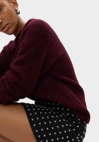 Bordeauxfarbenes, fuzzy Strickoberteil mit überschnittenen Schultern, kombiniert mit einem schwarzen Rock mit weißen Kreuzmustern. Nahaufnahme der Stofftexturen.