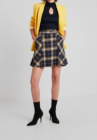 Blazer giallo sopra una maglia blu navy a coste con una fessura. Gonna plissettata a quadri in blu navy e giallo, abbinata a stivaletti neri alla caviglia.