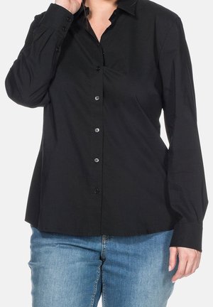 Henkilö pukeutuneena mustaan pitkähihaiseen napilliseen paitaan ja sinisiin farkkuihin, käsi kohotettuna lähellä kasvoja, yksinkertaista vaaleaa taustaa vasten.