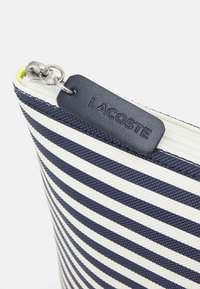 Pochette rayée bleu marine et blanche en tissu texturé, dotée d'une fermeture éclair et d'une étiquette en cuir bleu marine avec "LACOSTE" en relief.