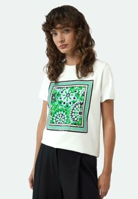 Witte katoen t-shirt met een kleurrijke bloemenprint in groen, blauw en zwart, omlijst door een geometrische zwarte rand en een geruit accent.