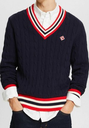 Hombre con suéter azul marino de punto grueso con escote en V y ribete a rayas en rojo, blanco y negro, sobre camisa blanca de botones, con las manos en los bolsillos.