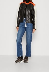 Žena v černé, bílé a oranžové kožené bundě, modrých džínech rovného střihu, černém roláku a hnědých teniskách stojí proti jednotné zdi.