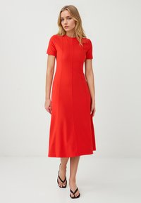 Vrouw in een aansluitende rode jurk met korte mouwen, staande voor een effen achtergrond, gecombineerd met zwarte flip-flops. Zelfverzekerde uitdrukking.