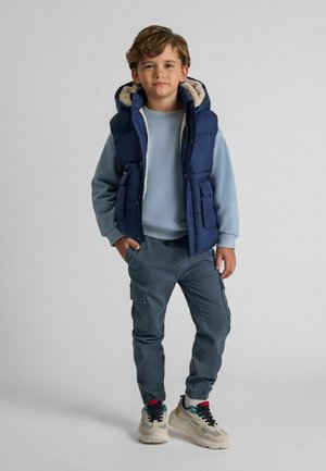 Gilet rembourré marine avec doublure en shearling, sweat-shirt bleu clair, pantalon cargo gris et baskets beiges avec accents colorés. Textures de tissu lisses.