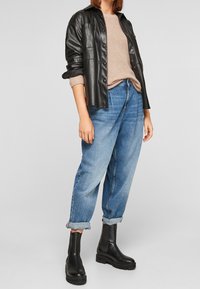 Czarna skórzana kurtka koszulowa na beżowym swetrze z dzianiny, w połączeniu z jasnoniebieskimi dżinsami i czarnymi botkami za kostkę. Zrolowane mankiety na dżinsach.