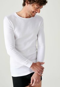 Langarmige witte thermoshirt met een geribbelde textuur, ronde hals en aansluitende mouwen, gedragen met zwarte shorts.