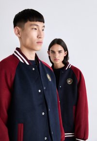 To modeller iført varsity jakker med mørkeblå kroppe og bordeauxfarvede ærmer. Hver jakke har en stripet ribbet krave og broderede logoer.