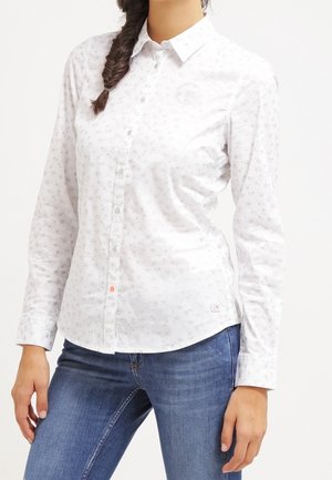 Vrouw draagt een wit, overhemd met lange mouwen en subtiele bloemenprint, gecombineerd met een blauwe spijkerbroek. Ze staat met één hand dicht bij haar heup.