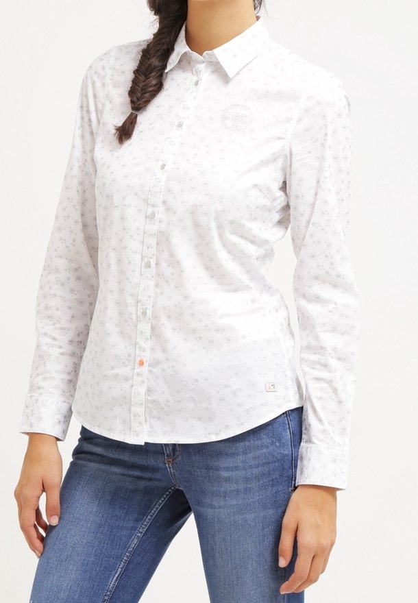 Femme portant une chemise blanche à manches longues avec des boutons, à imprimé floral discret, et un jean bleu, debout avec une main proche de la hanche.