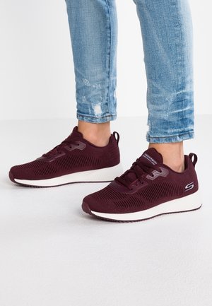 Persoon draagt lichtblauwe jeans met slijtage en bordeauxrode Skechers-sneakers met witte zolen, staand op een witte ondergrond.