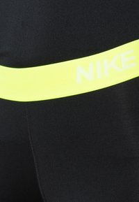 Svarta tights med en bred, klar gul midja som har en vit "NIKE"-logotyp. Materialet ser slätt ut med detaljer i sömnaden.
