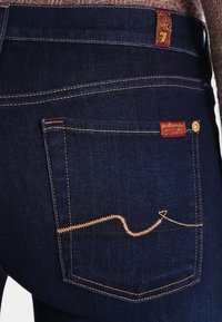 Jeans oscuros de mezclilla, con un bolsillo trasero que tiene un bordado naranja en un patrón de zigzag y una pequeña etiqueta roja encima del bolsillo.