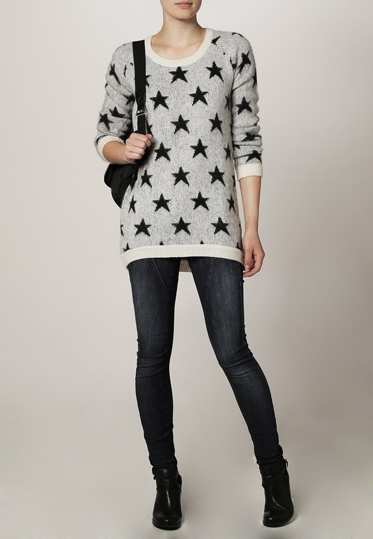 Personne portant un pull gris avec des étoiles noires, un jean skinny foncé, des bottines noires et tenant une sangle de sac à épaule noire.