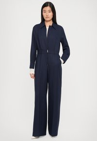 Námořnický overal s dlouhými rukávy, předním zipem a zúženým pasem; široké nohavice, hladká textura a minimalistické designové detaily.