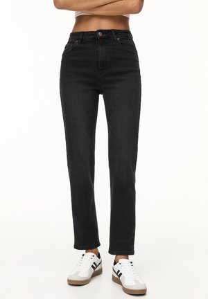 Personne portant un jean noir taille haute et des baskets blanches avec des bandes noires, debout les bras croisés sur un fond uni.