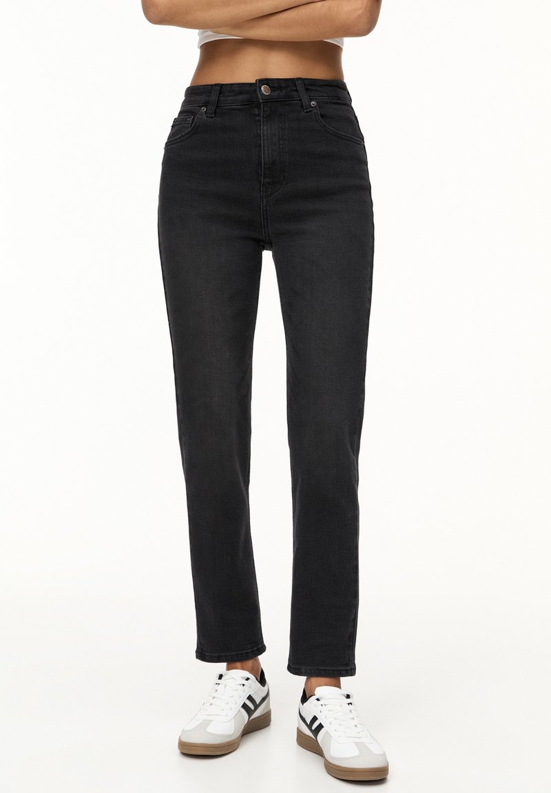 Personne portant un jean noir taille haute et des baskets blanches avec des bandes noires, debout les bras croisés sur un fond uni.