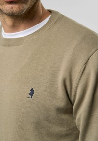 Beige pulóver kerek nyakkivágással és bordázott mandzsettával, amelyen egy kis sötétkék hímzett logó található a mellrészen. Puha tapintású anyag.
