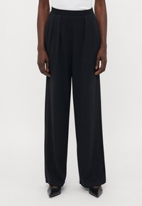 Schwarze weite Hose mit Falten, glatter Textur, hohem Bund und vorderem Bügel, kombiniert mit spitzen schwarzen Schuhen.