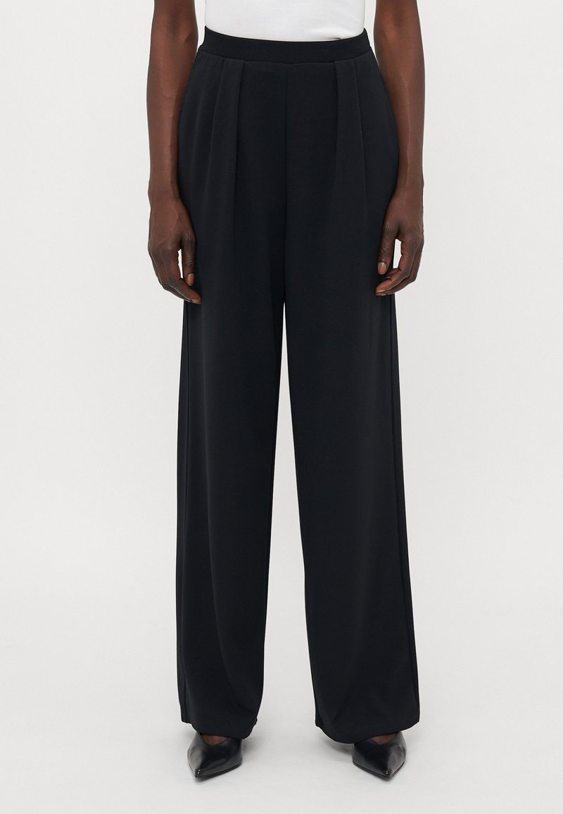 Schwarze weite Hose mit Falten, glatter Textur, hohem Bund und vorderem Bügel, kombiniert mit spitzen schwarzen Schuhen.