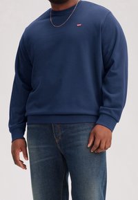 Uomo che indossa una felpa Levi's blu navy e jeans blu, con una sottile collana d'oro visibile su uno sfondo neutro.