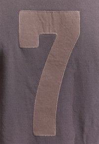 Grand patch en tissu beige en forme du chiffre « 7 » cousu sur un fond textile gris avec des coutures visibles autour des bords.