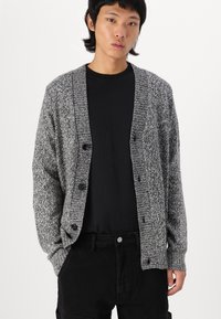 Grå strikket cardigan med teksturert finish, med knappelukking foran, lange ermer og en avslappet passform. Bært over en svart skjorte.