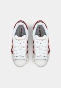 Zapatillas blancas con franjas rojas, presentando un upper de cuero suave, punta redonda y cordones planos. Acentos texturizados en la lengüeta y los laterales.