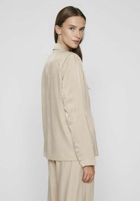 Blazer de tejido seda en color beige, con cuello, aberturas laterales y mangas largas, mostrado desde un ángulo lateral sobre un fondo neutro.