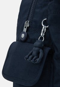 Läheltä otettu kuva tummansinisestä kangaskerrostöksestä, jossa on vetoketju, pyöreällä logolla varustettu vetopullo ja tekstuurinen apinamuotoinen avaimenperä, joka on kiinnitetty etutaskuun.