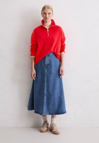 Rood fleece vest met ritssluiting gedragen over een beige coltrui, gecombineerd met een lange, blauwe spijkerbroekrok en beige sneakers met rode accenten.