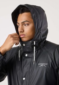 Czarna wodoodporna kurtka z kapturem, wyposażona w srebrne zatrzaski i regulowane troczki. Logo znajduje się po lewej stronie klatki piersiowej.