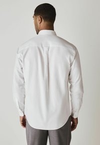 Weißes Langarmhemd mit einem geraden Kragen und einem Rückenfalten, kombiniert mit grauen Hosen. Der Stoff wirkt glatt und hat einen leichten Glanz.