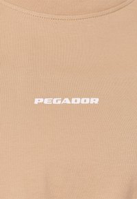 Beige tætsiddende t-shirt lavet af ribbet stof, med et hvidt "PEGADOR" logo i midten. Rund halsudskæring med syet detaljering.