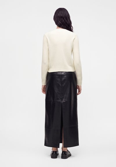 Femme aux longs cheveux foncés portant un pull crème et une longue jupe en cuir noire avec une fente centrale dans le dos, vue de dos.