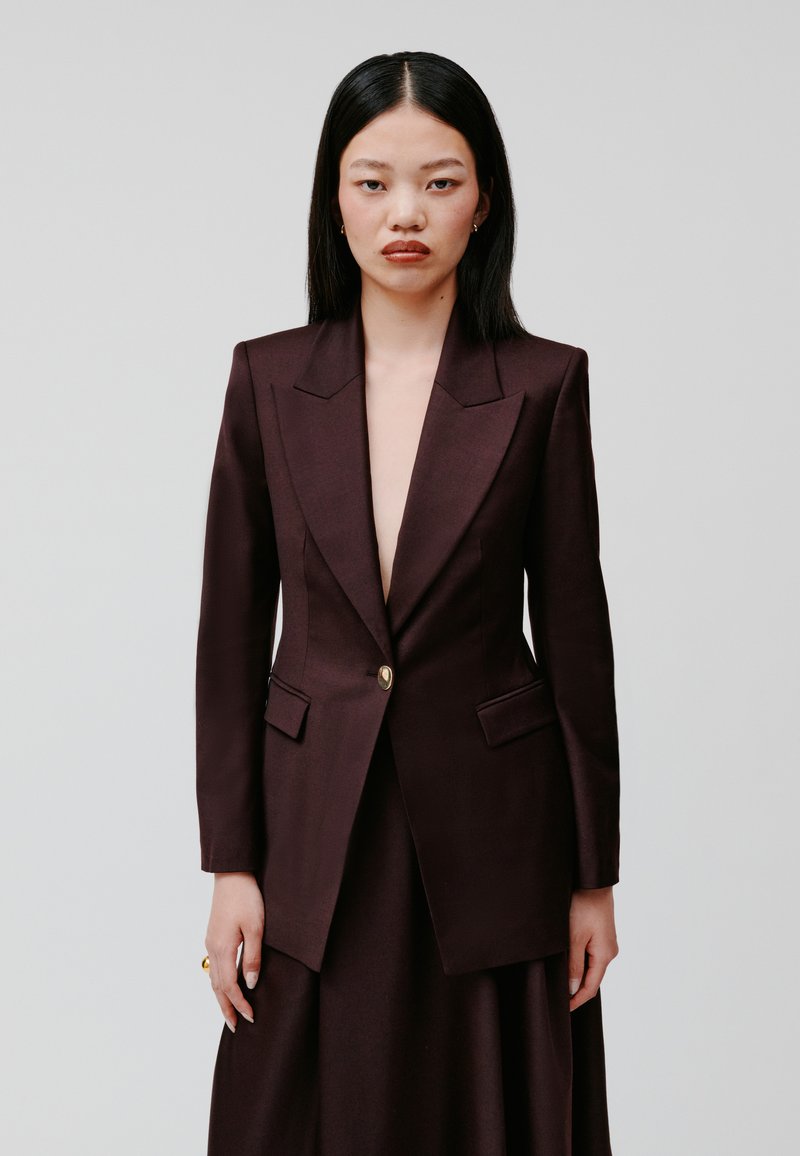 Burgundowa marynarka o dopasowanym kroju z głębokim dekoltem w literę V, wyraźnymi ramionami, złotymi guzikami i dwiema przednimi kieszeniami. Gładki materiał, nowoczesny krój.