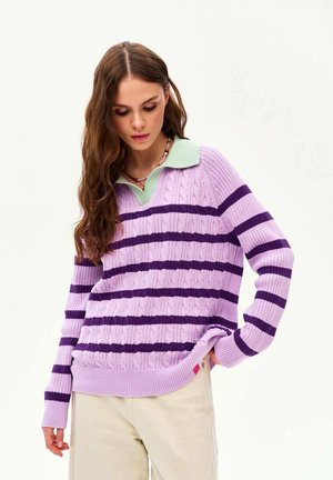 Kobieta o długich brązowych włosach nosi fioletowo-lawendowy sweter w paski z miętowym kołnierzem, w połączeniu z jasnobeżowymi spodniami, patrząc w dół.