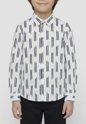 Bambino che indossa una camicia bianca a maniche lunghe con righe verticali blu navy e pantaloni neri, in piedi davanti a uno sfondo uniforme.
