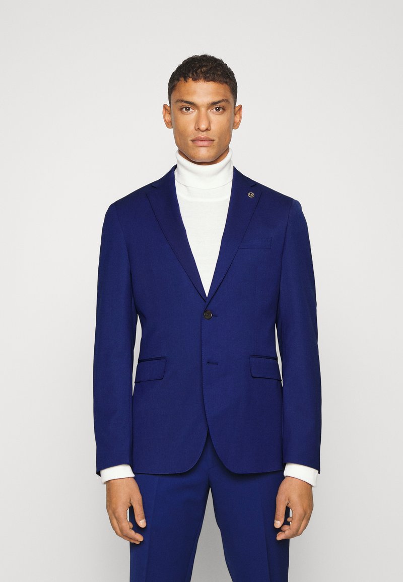 Marineblaues Sakk aus strukturiertem Stoff, mit einem einzelnen Knopfverschluss, Reverskragen und zwei vorderen Taschen. Darunter ein weißer Rollkragenpullover.