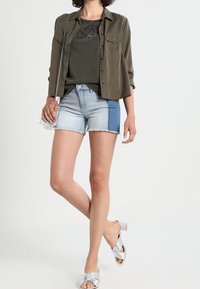 Olivově zelená košile na knoflíčky přes tmavé tričko s vyšitým designem, spárovaná s světlými denimovými šortkami se modrým bočním panelem. Stříbrné sandály.