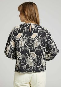 Blouse noire ornée de broderies florales et de feuilles blanches, avec des accents en dentelle et un ourlet festonné. Le dos présente une ouverture en forme de clé et un détail de bouton.